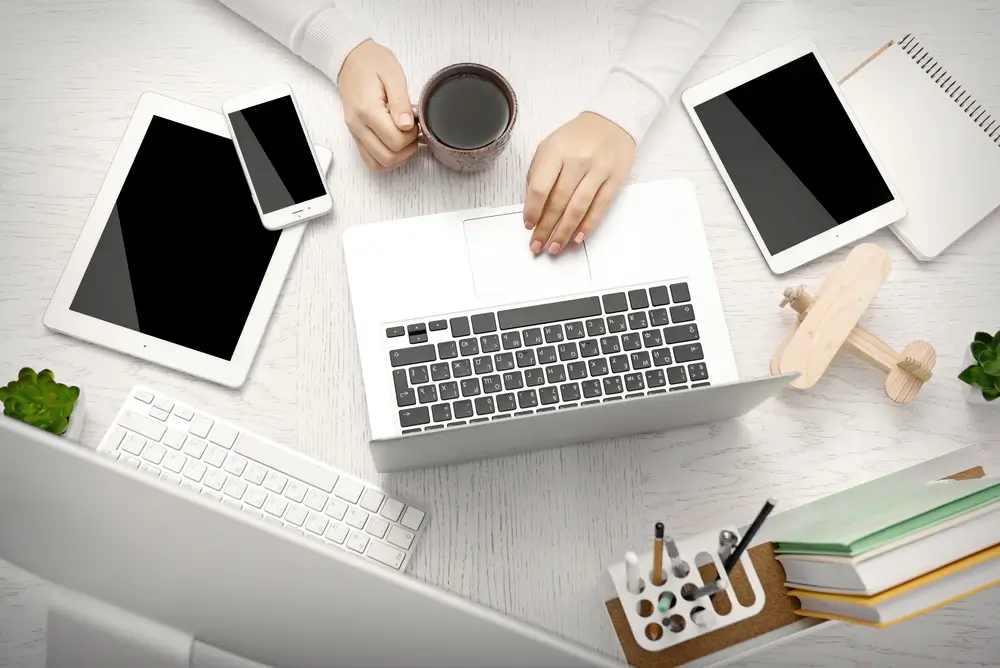 Kobieta korzystająca z laptopa przy biurku, na którym leżą telefon i tablet. Ubezpieczenie sprzętu elektronicznego.
